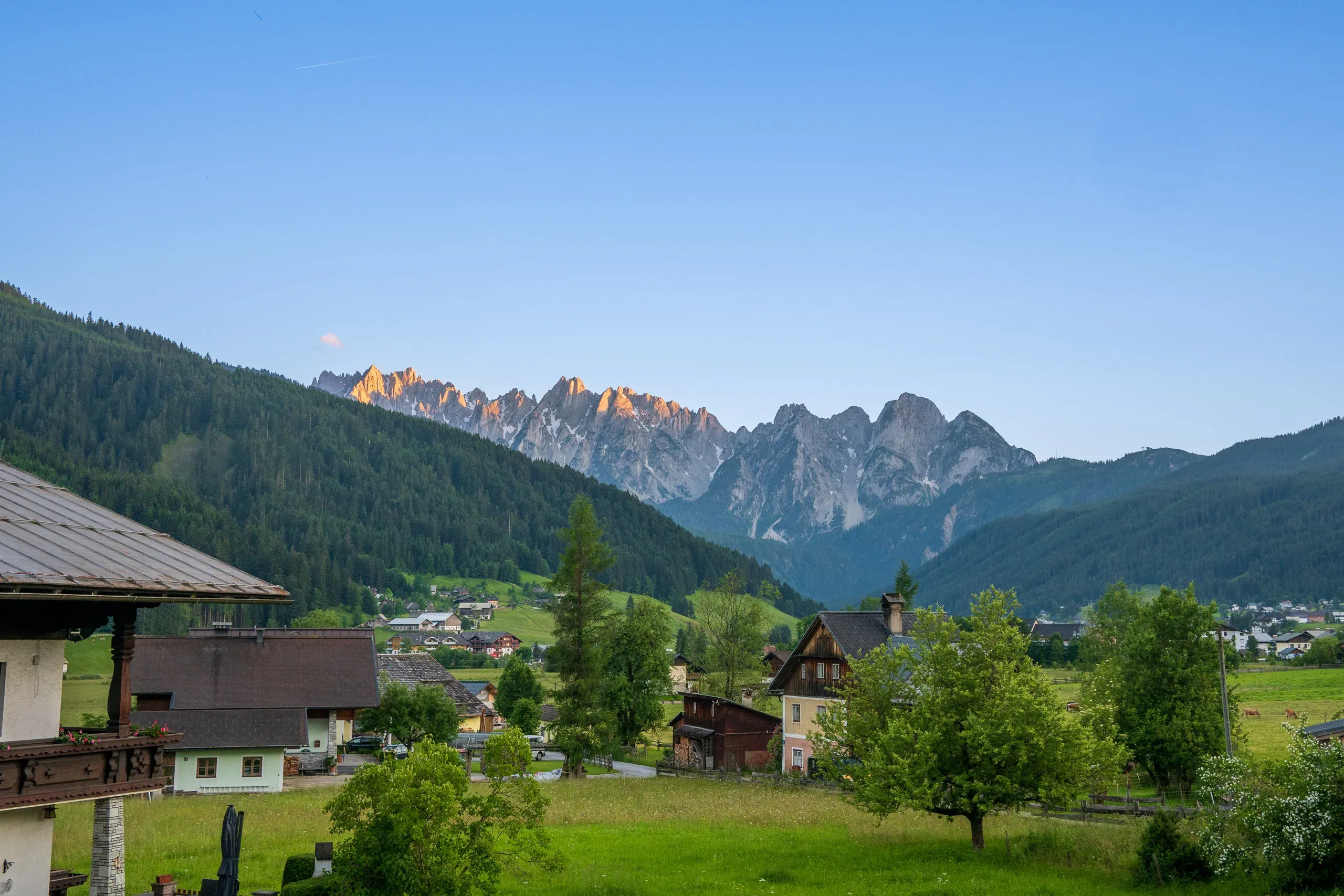 Alpendorf mit grünen Wiesen, dahinter felsige Gipfel im Morgenlicht