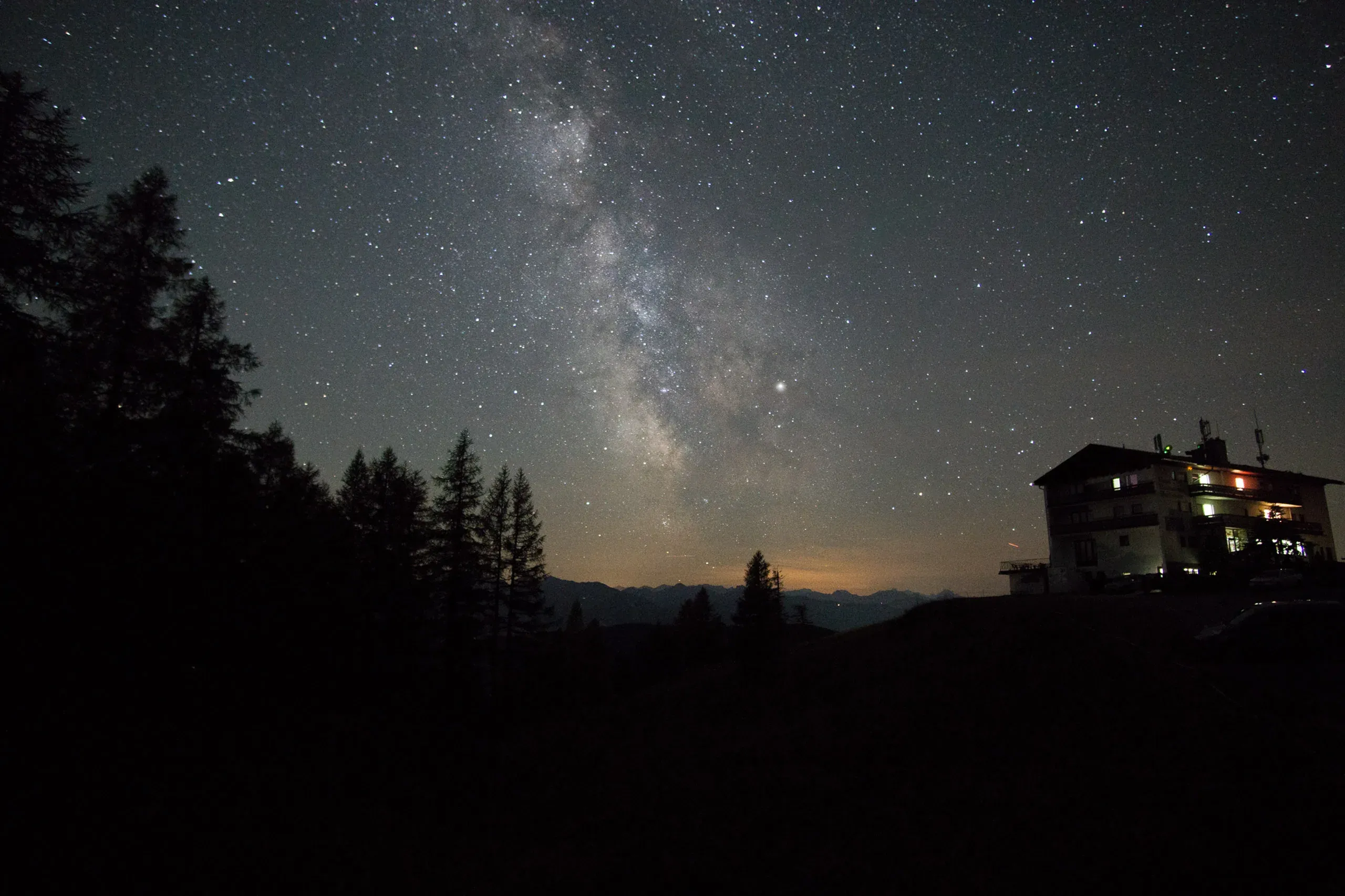 Milchstraße über einer beleuchteten Berghütte im Sternenhimmel